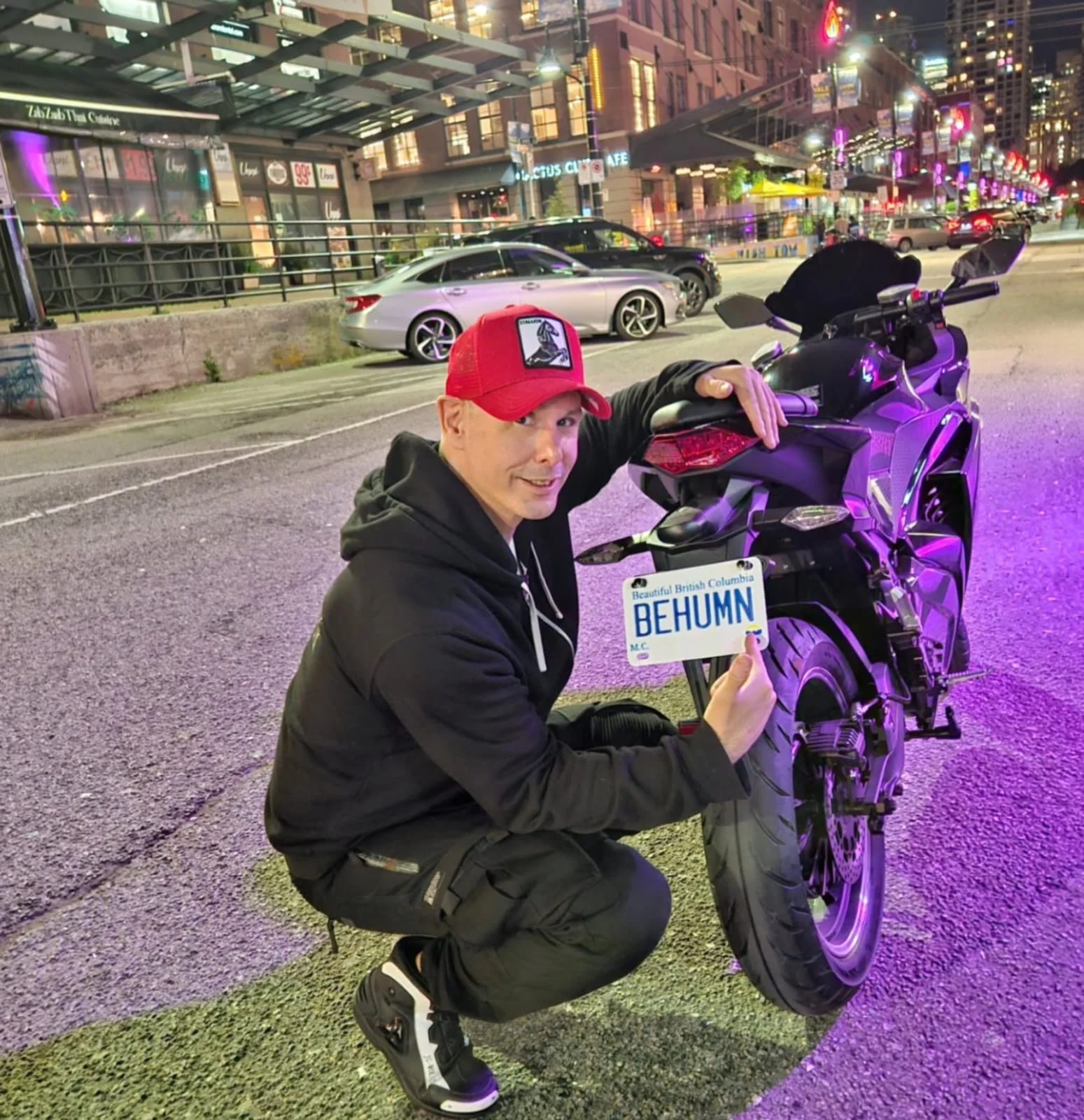 Shane James kneeling beside a motorcycle with a BEHUMN license plate, city lights behind him